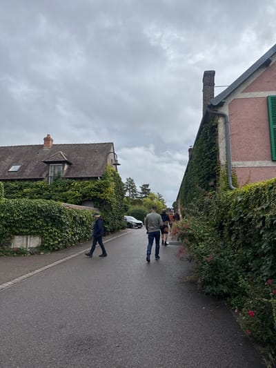 This image depicts a street scene in Giverny, France, under a heavily overcast sky. The asphalt road, appearing damp, extends into the background.

On the left side of the street, a two-story building with a brown-tiled gabled roof features a central dormer window, a skylight, and a red brick chimney. The building's walls and a low retaining wall extending from it are densely covered in green ivy. A black, curved street lamp protrudes from the ivy-covered wall. A rectangular, light-colored utility box is affixed to the lower section of this wall. Further down the street on the left, a dark-colored sedan is parked.

On the right side of the street, a two-story building with a light pink stucco facade has its visible side wall extensively covered in green ivy. A grey downspout runs vertically along this wall. The dark roofline and a brick chimney are visible. A partially obscured window with closed green shutters is visible on the upper right portion of the building.

Several individuals are present on the road. In the foreground on the left, a person wearing a dark blue jacket, dark trousers, and a light-colored cap walks towards the left side of the frame. In the mid-ground, a male figure wearing a light grey long-sleeved shirt and blue jeans walks away from the viewer. Behind him, a second individual in dark clothing also walks away. Multiple indistinct figures are visible further down the street, also moving away from the viewer.