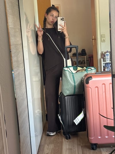 The image shows a young woman with braided hair taking a mirror selfie in what appears to be a domestic setting. She is wearing a dark brown jumpsuit and a white crossbody bag. In front of her are two suitcases, one black and one pink. The background reveals a hallway with a door, shelves with items, and a power outlet on the wall. The overall impression is that of someone preparing to travel. The specific location within Bruchköbel, Germany, is not determinable from the visual cues provided in the image.