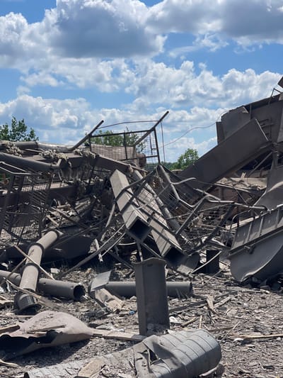 The image shows a scene of destruction in Terny, Ukraine. The location is a site of a destroyed building, likely an industrial facility, with rubble scattered across the ground. The site contains large pieces of metal, twisted beams, and other debris, suggesting an explosive event. There are no visible people in the image. The debris is lying on a field of dry, cracked earth and dirt, with green foliage in the background. The sky is blue with white clouds.