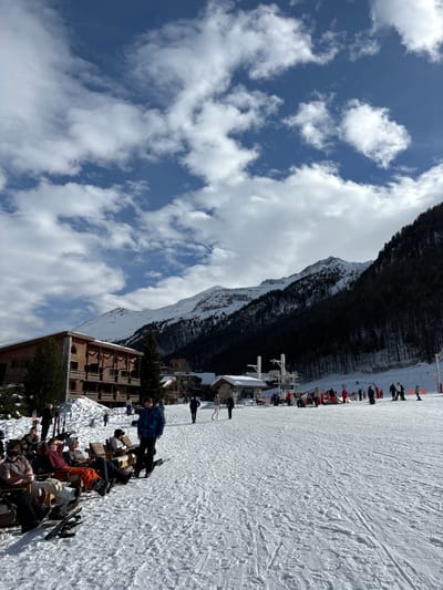 This image depicts a vibrant outdoor scene at a ski resort, likely Val-d'Isère, France, under a clear blue sky dotted with scattered white cumulus clouds. The photograph, originally captured in a portrait orientation but showing a landscape scene, reveals a broad ski slope filled with tracks, leading up towards snow-capped mountains and a dark, dense forest in the background.
In the mid-ground, at the base of the slope, are resort buildings and infrastructure. Notably, a prominent white sign with black lettering reads "LA FOLIE DOUCE," identifying a famous après-ski establishment. Next to it are other white structures, possibly related to ski lift operations.
The foreground is bustling with activity, showing numerous people gathered at an outdoor seating area. Many individuals, dressed in colorful ski jackets, pants, and helmets, are either seated at tables or standing, socializing and relaxing. Skis and poles are visible, propped against tables or lying in the snow. A multi-story wooden lodge-style building is situated on the left side of this gathering area. The time of day appears to be midday or early afternoon, with bright sunshine casting distinct shadows, indicating good weather conditions. The overall atmosphere is relaxed and social, suggesting a break from skiing or an après-ski event.