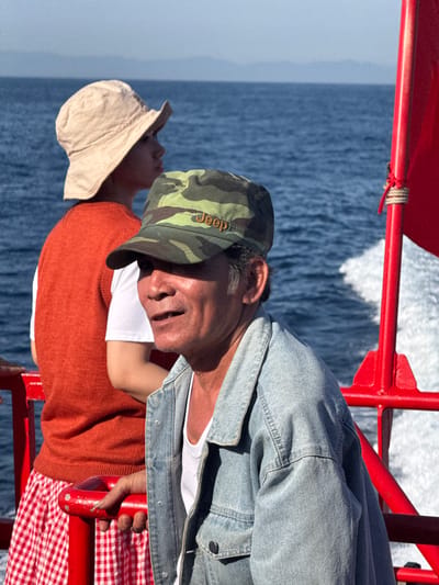 Two people are on a boat on the sea. The person in the foreground is a man wearing a denim jacket and a camouflage cap. He is looking to the right with his mouth open. The person behind him is wearing a cream-colored hat and an orange sweater. They are looking to the left. The water is blue and there is white wake behind the boat. A red railing is visible in the foreground. The image was taken in the city of Dong Son, Vietnam.