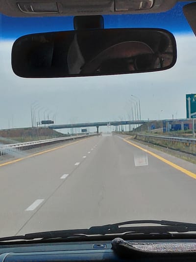 The image depicts a view from inside a vehicle, looking forward through the windshield and up at the rearview mirror. The rearview mirror reflects the driver and a passenger wearing sunglasses. The vehicle is on a highway, with multiple lanes and a yellow dividing line. Overhead structures, possibly bridges or overpasses, are visible in the distance, along with streetlights and signage. The sky is overcast. The scene is consistent with a highway setting in a city, potentially Aqsu city administration, Kazakhstan, given the context provided.