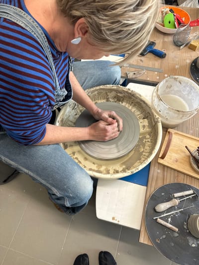 This image captures an individual engaged in pottery work within a studio in Mafra, Portugal. The scene, viewed from an overhead perspective, shows a person with short blonde hair wearing a blue and maroon striped shirt under light-wash denim overalls. Their hands are clasped over a lump of grey clay centered on the spinning wheel head of a pottery wheel, indicating the process of throwing. A white basin on the pottery wheel collects water and clay slurry.
To the right, a light-colored wooden workbench is visible, cluttered with pottery tools and materials. These include a white plastic bucket containing a pale liquid (likely slip or water), a rectangular wooden tray with a brown leather handle marked "NORWOOD" holding small implements, a blue-handled rubber spatula, a metal ruler, a yellow sponge, and various other metal and wooden pottery tools. Further right, a circular dark surface displays a small, unglazed clay pot and additional tools, including a loop tool with a wooden handle. The person's feet, clad in black split-toe shoes, rest on a light grey tiled floor in the foreground.