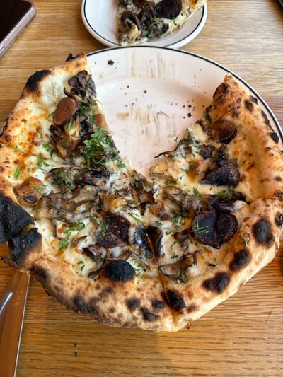 The image shows a partially eaten pizza on a white plate with a black rim, sitting on a wooden table. A slice of pizza is on a smaller plate with a black rim to the left of the main plate. The pizza appears to be a mushroom pizza, with various types of mushrooms, cheese, and parsley as toppings. The crust is charred in places. A knife and a cellphone are visible near the bottom of the image. The location is Culver City, United States of America.