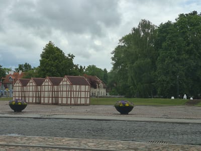 The image depicts a town square with cobblestone paving and several buildings and natural elements. In the foreground, a cobblestone street leads the eye towards a plaza area. Two large, dark, bowl-shaped planters filled with purple and yellow flowers are positioned on the plaza. Behind the planters, a row of four half-timbered structures with red roofs stand prominently. These structures appear to be temporary or decorative, possibly market stalls or exhibition booths, given their uniform design. To the left of these structures, a traditional building with a red tiled roof and white walls is visible, alongside a taller, lighter-colored building with multiple windows and a flag pole. The background is dominated by lush green trees, suggesting a park or well-maintained green space. The sky is overcast with a cloudy, grey tone. The scene is quiet and appears to be unoccupied by people. The architecture of the half-timbered buildings and the traditional house suggest a European setting. This scene is located in Šilutė, Lithuania, specifically in the area around the town's central square or a public gathering space.