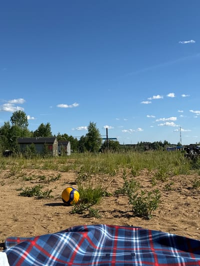 The image depicts an outdoor scene on a sunny day in Рыбхоз, Russia. In the foreground, a blue, red, and white checkered blanket is spread on sandy ground. A yellow and blue volleyball rests on the sand to the left of the blanket. The background features dry, sparse vegetation and a line of trees. Several small, grey structures with black roofs and red doors are visible among the trees. Further in the distance, industrial structures and a communication tower can be seen. The sky is clear blue with scattered white clouds.
