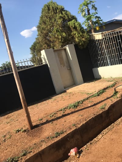 The scene is located in the Mandate Estate in Ilorin, Nigeria. The image depicts a building with a metal-barred window and a metal door. A large tree stands next to the building, reaching up into a partly cloudy sky. A wooden pole extends across the foreground. The ground is dirt and gravel, with some vegetation. A piece of trash lies on the ground.