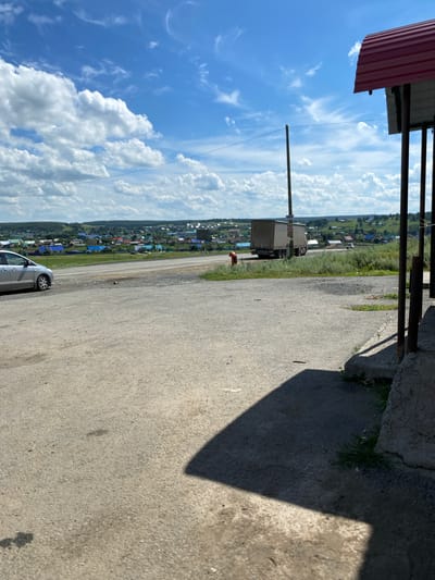 A bright, sunny day is depicted in Yuldybayevo, Russia, with a clear blue sky dotted with fluffy white clouds. In the foreground, a paved area transitions into a grassy verge. A silver car is parked on the left side of the paved area, facing left. Beyond the car, a road runs horizontally across the frame. In the middle ground, a large, beige truck is driving away from the camera on the road. Further in the distance, a cluster of houses with colorful roofs spreads across a gently rolling, green landscape, indicative of the village of Yuldybayevo. The right side of the image shows the corner of a building with a red metal roof and dark vertical supports. The overall scene is a rural roadside view with a village in the background.