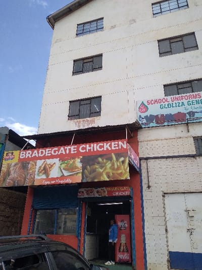 This image captures a street view of a multi-story commercial building during the daytime. The building's exterior is painted a light cream or white color, with several windows visible on its upper floors. These windows have dark frames and appear to be fitted with security bars or a grid pattern.
On the upper part of the building, a large horizontal banner is displayed, advertising a business named "SCHOOL UNIFORMS GLOLIZA GEN". Below this text, there is a logo featuring the number "25" within a stylized shape, and possibly additional text that is difficult to read.
The ground floor of the building houses commercial establishments. On the left, a prominent vertical banner advertises "BRADEGATE CHICKEN". This banner showcases appetizing images of fried chicken, french fries, and other chicken dishes. Below these images, the phrase "Enjoy our Hospitality" is printed.
Adjacent to the large "BRADEGATE CHICKEN" banner, to the right, is the entrance to a shop, presumably also part of "BRADEGATE CHICKEN", as a smaller sign above its entrance also reads "BRADEGATE CHICKEN". The walls around this entrance are painted in bold red and blue sections. Inside the entrance, a Coca-Cola branded refrigerator is visible, and a person wearing a blue top is standing near it.
Part of a dark-colored vehicle, possibly black, is visible on the far right edge of the image, suggesting it is parked on the street in front of the shops. The sky in the background, visible in the upper left, is bright blue with some light clouds, indicating clear weather during daylight hours. The overall scene suggests a busy commercial area.