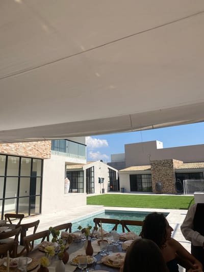 The image shows an outdoor dining area next to a swimming pool. The setting appears to be residential, with multiple modern houses visible in the background. A white canopy stretches overhead, providing shade. A long dining table is set for a meal, with glassware, plates, and floral arrangements. Several people are present, primarily around the table. The location is likely in Santa Cruz Tehuixpango, Mexico, based on information provided.