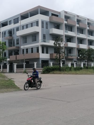 An outdoor, eye-level shot captures a modern residential or commercial area in Ha Tu Ward, Vietnam, on an overcast day. In the foreground, a man sits on a red and black motorbike on a concrete road. He wears a blue long-sleeved shirt, dark pants, and a dark helmet, appearing to be looking down at something near his hands. To the left of the road, there are patches of dry grass.
The background features a large, multi-story building with a contemporary design, characterized by a mix of light gray, white, and light brown architectural elements. It has numerous dark-framed windows and balconies with glass railings. The top floor appears to be an open-air structure. A white-pillared fence with dark metal railings runs along the base of the building, separating it from the road. Scattered green bushes and small trees are visible along the fence line and adjacent to the building. The sky is uniformly gray, indicating an overcast day. No discernible text is visible in the image.