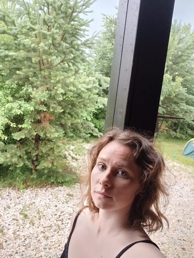 A medium shot captured from a low angle shows a woman with wavy brown hair and light skin looking directly at the camera. She is positioned in the foreground, slightly off-center to the right, with a dark-colored garment visible on her shoulders. The background features lush green trees, a dark-colored vertical structure with water droplets on its surface, a gravel-covered ground, and a portion of a green tent in the distance. The overall scene suggests an outdoor setting, possibly in Verkhneye Dubrovo, Russia, on a cloudy or rainy day.