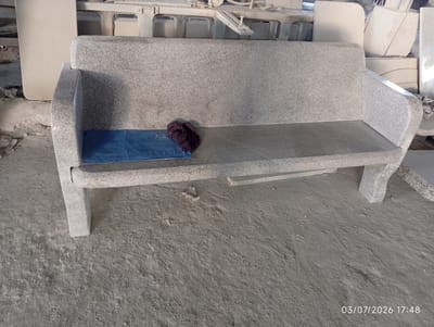 In the city of Bana Stream, Nigeria, a finished granite bench is displayed indoors. The bench is light gray with speckles and has a solid, sturdy construction. A blue cloth is placed on the seat of the bench, and on top of the cloth, a small, dark purple knitted item rests. The bench is situated on a concrete floor within what appears to be a workshop or showroom, indicated by the presence of other stone materials and industrial equipment in the background. The lighting suggests an indoor environment, possibly a factory or storage area. The timestamp "03/07/2026 17:48" is visible in the lower right corner of the image.