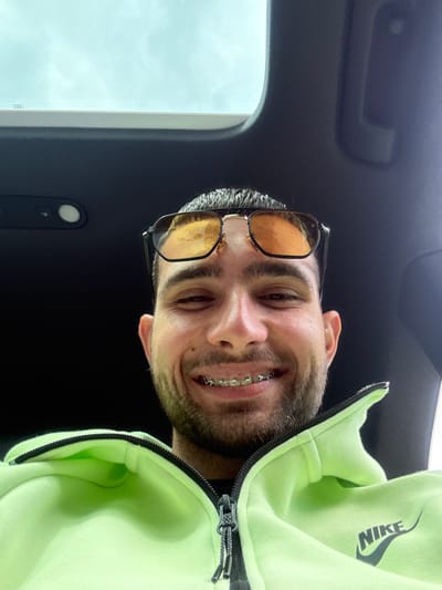 A man with glasses and braces smiles at the camera. He is wearing a bright green Nike sweatshirt. The image appears to be taken from inside a vehicle, possibly an Audi, judging by interior features. The background shows the car's interior and a glimpse of the outside, revealing sky. There is no evidence indicating the location to be Dolna Studena, Bulgaria.