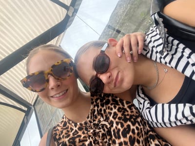 The image depicts two young adult females under a striped canopy structure, consistent with a bus stop, in Ribamontán al Mar, Spain. Both subjects are positioned closely together, facing the camera in a selfie-style perspective.
The female on the left is smiling, wearing large, tortoise-shell, cat-eye sunglasses and a leopard print top. Her light-colored hair is pulled back.
The female on the right has her head resting on the left female's shoulder. She wears dark-rimmed sunglasses, a black tank top, and a black and white horizontally striped cardigan. A silver-toned, double-layered necklace, one strand with a small star pendant, is visible on her neck. Her fingernails feature a French manicure with small pink star embellishments on her ring finger. A black bag with silver stud detailing is partially visible on her right.
The immediate environment includes the overhead striped canopy, supported by dark beams. Beyond the subjects, through a clear panel, green foliage and an overcast sky are visible. A white sign with black text, including the acronym "TUS," is partially discernible behind the left female, indicating a public transportation information board.