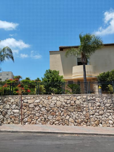 The image depicts a street view in Zikhron Yaakov, Israel, on a bright, sunny day with a clear blue sky and scattered white clouds. In the foreground, a paved road runs along the bottom of the frame, with a sidewalk made of brick pavers and a concrete curb separating it from a large, rough-hewn stone wall. This wall, constructed from a variety of natural stones in shades of tan, brown, and gray, forms the base for the property behind it. A thin, reddish-brown pole extends vertically from the sidewalk to the top of the stone wall, with a small, yellow sign attached to it.
Behind the stone wall, a black metal fence runs horizontally, enclosing a garden area. This garden features lush green foliage, including trees and flowering bushes with vibrant red blossoms. A taller, modern building with a cream-colored facade and a distinctive curved balcony with square cutouts is visible behind the garden. A palm tree with a slender trunk and a full canopy of fronds grows in front of this building, partially obscuring a window. To the left of this building, another, taller apartment building with a lighter facade is partially visible. To the right of the cream-colored building, more dense green vegetation and another palm tree are present. A yellow propane tank is visible next to the fence on the right side of the image. The overall scene is one of a residential area, likely a street in Zikhron Yaakov, characterized by natural stone construction and Mediterranean vegetation.
