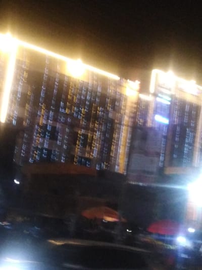 A blurry, low-light image shows a building at night adorned with strings of lights, creating a festive atmosphere. The lights appear as streaks of yellow and blue. Below the building, two red umbrellas stand out amidst the darkness and other indistinct shapes, potentially indicating a street market or gathering. The overall image is hazy, likely due to motion blur or a poorly focused camera.