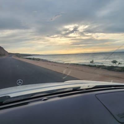 The image is taken from the interior of a vehicle driving along a coastal road. The road is paved and curves to the left. To the right of the road is a sandy embankment leading to a body of water, likely a lake or sea. The water is calm with gentle waves. The sky is overcast with patches of sunlight breaking through, creating a soft, diffused light. The landscape is arid with sparse vegetation along the shore and distant mountains visible on the horizon. A prominent Mercedes-Benz hood ornament is visible in the foreground, indicating the type of vehicle. The overall impression is one of a scenic drive along a remote coastline in a desert or semi-arid environment. The location is identified as the city of Tosor, Kyrgyzstan.