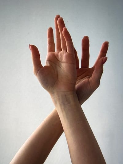 This is a close-up, medium shot photograph of two human hands, with their arms visible in the foreground. The hands are positioned in the upper portion of the frame against a plain, light gray background. The left hand is angled slightly upward and to the right, with its palm facing the viewer. Its fingers are spread open, with the thumb extended. The right hand is positioned behind and to the right of the left hand, with its palm facing away from the viewer and angled slightly downward. Its fingers are also extended, with some overlapping the fingers of the left hand. The forearms of both individuals are visible in the lower half of the image, crossing each other. The skin tones are fair, and the fingernails on the visible fingers appear to be painted with a light pink or nude polish. The lighting is soft and diffused, creating subtle shadows that highlight the contours of the hands and arms. There are no discernible elements in the background that suggest a specific location or environment. The image does not contain any clues that can be used to determine its location in the city of Genemuiden, Netherlands.