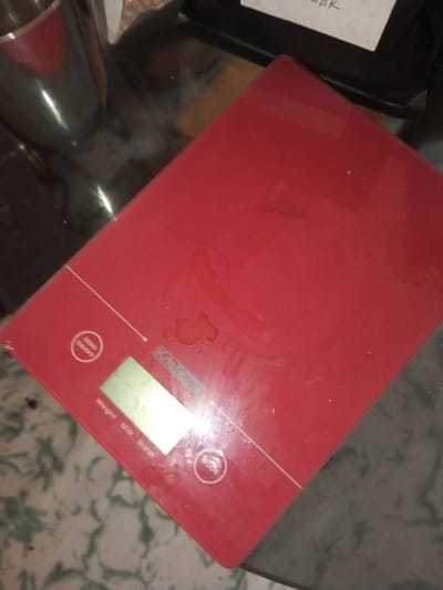 A red digital kitchen scale sits on a reflective surface. It's rectangular with rounded corners and shows buttons labeled "zero/on/off", "Weight Milk Water", and "Mode". The display screen is visible. The brand name "Kris Chef" is printed on the scale. There are several splotches and streaks of liquid on the surface, suggesting spills. A stainless steel object sits to the left of the scale, and a bag with writing on it is to the upper right.