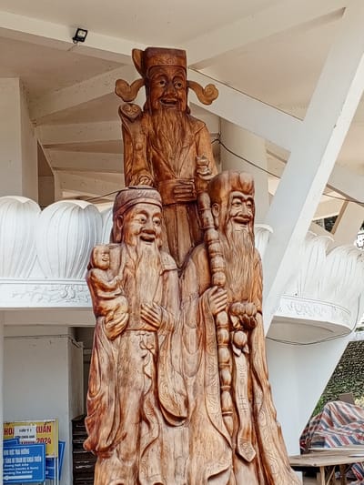 A close-up, low-angle shot captures a wooden carving of three elderly men with long beards, possibly representing deities or historical figures from East Asian culture. The central figure stands tallest, wearing a headdress and robes, with a benevolent expression. To his left, a figure holds a baby and a staff, also smiling. To the right, another figure grasps a staff and holds a cluster of fruit or similar objects, with a wide grin. All three figures are depicted in flowing robes with intricate detailing.
In the background, a white, spiral staircase is visible, partially obscured by the figures. The architecture suggests an outdoor covered area, possibly a temple or public space. White decorative elements, resembling lotus petals, are present on a ledge below the staircase. A sign with Vietnamese text is visible in the lower left corner, indicating a location in Nam Ban, Vietnam. The overall lighting suggests daylight.