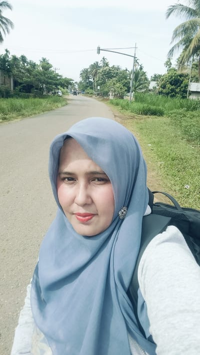 A selfie shows a woman wearing a light blue hijab and a gray sweatshirt. She is standing on an asphalt road with greenery and palm trees on either side. In the background, a traffic light and several vehicles are visible. The sky is bright and cloudy.
