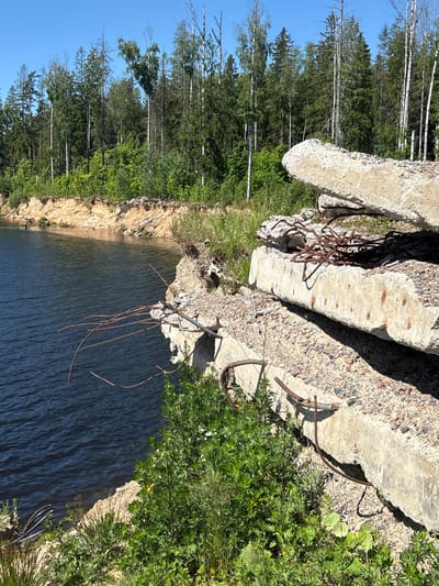 The image depicts a disused quarry in Yoksolovo, Russia, characterized by a body of dark blue water on the left and a steep, sandy embankment leading up to a dense forest of coniferous and deciduous trees under a clear blue sky. In the foreground on the right, a pile of broken concrete slabs is prominently featured, with exposed rusted rebar protruding from the debris. The slabs appear to be remnants of construction or demolition. Lush green vegetation, including bushes and smaller plants, grows around the concrete rubble and along the edge of the water. The overall scene suggests a state of abandonment and natural reclamation.
