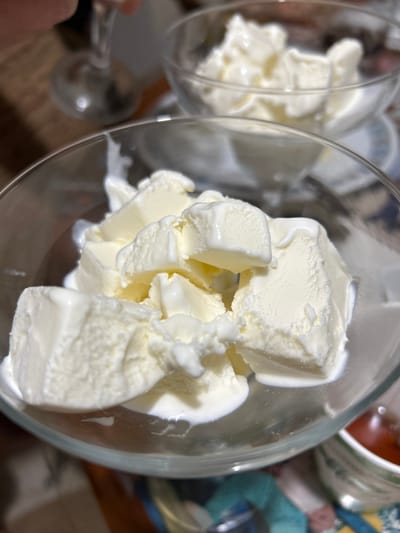 A close-up shot shows two glass bowls filled with scoops of melting vanilla ice cream. The bowl in the foreground is larger and fills most of the frame, showcasing creamy, soft ice cream with a slight sheen of melt. The bowl in the background is smaller and out of focus, also containing scoops of ice cream. The setting appears to be indoors, with a wooden table and a blurry background that suggests a dining area. The image does not provide information about the city of Copceac, Moldova, or any specific details about people or actions occurring within the scene.