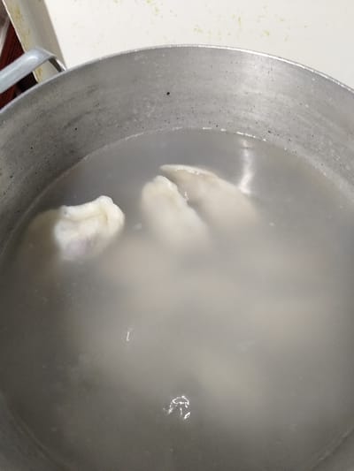 A close-up shot shows several dumplings submerged in cloudy water within a silver pot. The dumplings appear pale and somewhat translucent, with one on the left showing a slight purplish hue. The pot itself is made of metal and shows some signs of wear. The water is murky and appears to be heated, as evidenced by a few small bubbles and wisps of steam. The background is out of focus but suggests a light-colored surface, possibly a countertop. The scene is set in the city of Shchastia, Ukraine, within what appears to be a domestic kitchen environment.
