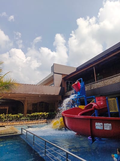 A colorful water park structure, featuring a red bucket and slides, stands beside a pool. Water gushes from the bucket onto the slides. A metal railing surrounds the pool. Lush greenery lines the edge of the pool near a low building with arched entrances. The architecture includes a light-colored building extension above. A sky with scattered clouds is visible above the structures.
