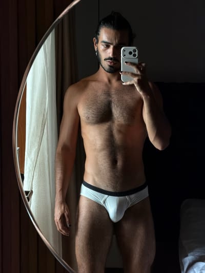 A man is captured in a mirror selfie, wearing white briefs and holding a smartphone. The background suggests an indoor setting, possibly a bedroom, with curtains visible on the left and a dark wall or furniture on the right. The image does not contain any identifiable landmarks or elements that would place it specifically in Pasir Putih, Indonesia.