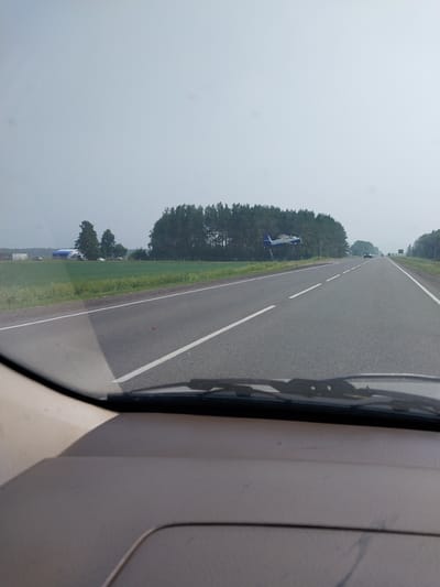 This is a photo taken from inside a vehicle, looking out the windshield. The view is dominated by a road stretching into the distance, bordered by green fields and a dense forest on the left. The sky is overcast, suggesting a cloudy or hazy day.

On the left side of the road, a blue and white structure, possibly an airplane or a model of one, is visible mounted on a stand. Further down the road, several vehicles are seen in the distance, traveling in the same direction. The right side of the image shows the interior of the vehicle, including the dashboard and part of the windshield frame.

The scene appears to be a rural or semi-rural setting, possibly in Belokurikha, Russia, given the provided location context. The time of day is unclear due to the overcast conditions. There is no visible text in the image. The overall mood is neutral, conveying a sense of travel or transit.