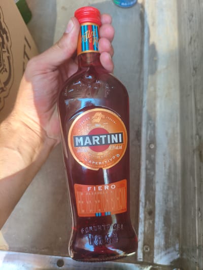 An overhead, slightly angled shot captures a hand holding a bottle of Martini Fiero vermouth. The bottle, predominantly red with an orange label featuring the Martini logo and text, is held in the center of the frame. The label text includes "MARTINI," "APERITIVO," and "FIERO," along with "11.9% alc. vol." and some Cyrillic text reading "НЕ ЯВЛЯЕТСЯ ВИНОМ" (not a wine). The hand is positioned around the neck and upper body of the bottle. In the background, a blurry, corrugated metal surface is visible, suggesting an outdoor or industrial setting. There is no visible indication of the city of Alchevsk, Ukraine, in the image.