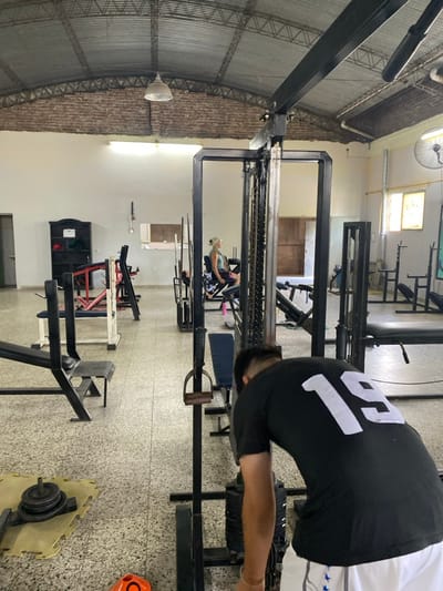 A gym features various weight machines and equipment. A man wearing a black shirt with "18" on the back is leaning over one of the machines. A woman is visible in a mirror on a weight machine. The gym has high ceilings and a tiled floor with diverse workout stations.