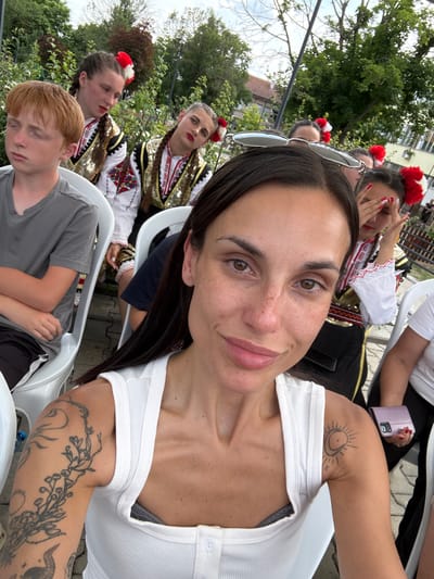 In Pordim, Bulgaria, a group of people are seated outdoors, likely attending an event. In the foreground, a woman with dark hair and freckles is taking a selfie, smiling at the camera. She is wearing a white tank top and has tattoos on her left arm. Behind her, a young boy with red hair is looking down, and several girls in traditional Bulgarian folk costumes, adorned with red and white pom-poms, are also visible. The background shows trees and buildings, suggesting an urban or semi-urban setting.