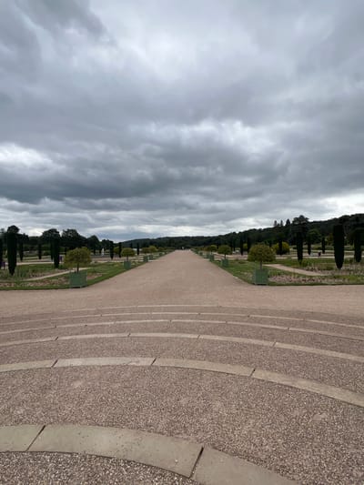 The image depicts a long, wide gravel pathway leading into a verdant garden landscape. The pathway is flanked by manicured trees and shrubs, some of which are contained in green square planters. In the distance, a fountain is visible, and beyond that, a line of dense green trees forms the horizon. The sky above is overcast with heavy grey clouds. In the foreground, there are curved, tiered sections of gravel, appearing to be part of a grand entrance or viewing area. The overall impression is of a formal, expansive garden setting. The location is not Stafford, United Kingdom; it is the Gardens of Versailles, France.