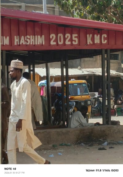 The image depicts an outdoor street scene in Tudun Wazurchi, Nigeria, under bright daylight. A prominent red metal-framed shelter, likely a bus stop or market stall, stands in the midground, its fascia displaying white lettering that reads "IM HASHIM 2025 KMC". In the left foreground, a dark-skinned man, wearing a light-colored traditional tunic and trousers with a patterned round cap, walks from left to right, partially obscured. Beneath the shelter, a yellow three-wheeled auto-rickshaw is parked, accompanied by several motorcycles. Another individual, dressed in light-colored attire, is seated on the ground under the shelter, facing away from the camera. The ground in the foreground is unpaved earth, scattered with various pieces of litter, including plastic bags. In the background, a cluster of buildings with corrugated metal roofs and more distant multi-story concrete structures are visible, interspersed with green foliage.