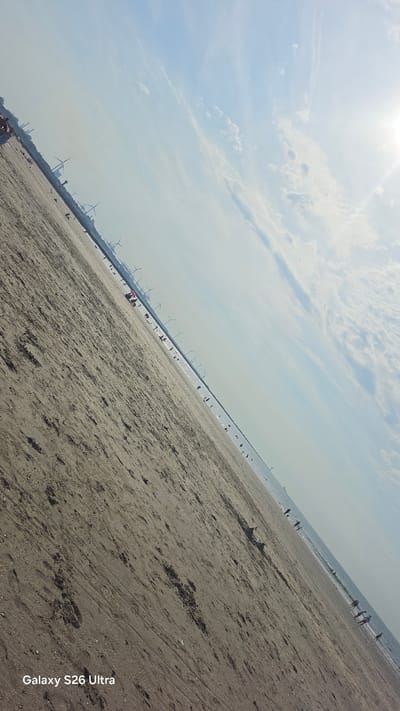 This media file depicts a wide, sandy beach with several people enjoying a day by the sea. The scene is set at Hook of Holland, Netherlands, with numerous wind turbines visible along the coastline, suggesting a coastal area with renewable energy infrastructure. The sun is high in the sky, casting a bright light and indicating it is daytime, likely late morning or early afternoon. The sky is partly cloudy with wispy clouds scattered across a pale blue expanse.

Several individuals and small groups are scattered across the beach and along the water's edge, some walking, others appearing to be relaxing or playing. The water is calm and a muted blue, meeting the sandy shore where small waves are lapping. The sand itself appears somewhat damp and textured, with evidence of footprints and debris.

Further in the distance, a long pier or breakwater stretches out into the sea, lined with more wind turbines. A few more people can be seen walking along this structure. The overall atmosphere seems relaxed and pleasant, indicative of a typical beach outing.

The visible text in the bottom right corner reads "Galaxy S26 Ultra," which is likely a watermark from the device used to capture the image. There are no other discernible text elements within the scene itself. The image is captured at a diagonal angle, adding a dynamic perspective to the composition.