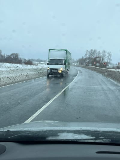 A white GAZelle-type light truck with a high, green box body is observed driving on a wet, asphalt road, seen from the perspective of a vehicle's dashboard and windshield. The truck's headlights are illuminated, and its Russian license plate, "Х584КХ 98," is visible. The road surface is visibly wet from precipitation, with a solid white line delineating the left side of the lane. The road curves sharply to the right in the mid-ground, indicated by red and white chevron signs positioned on the right shoulder. Snow-covered verges line both sides of the road, and the background features sparse, leafless trees and shrubs under an overcast, light grey sky. This scene occurs on a road in Rogachovo, Russia, during cold and damp weather conditions.