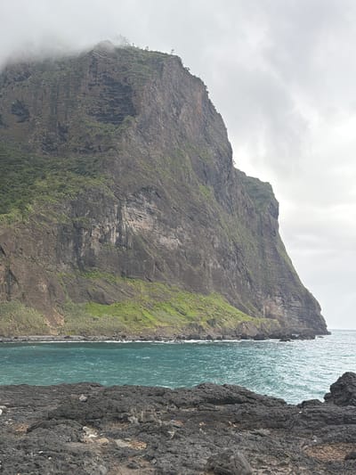 The image shows a rugged, rocky coastline adjacent to the ocean. A large, sheer cliff face dominates the left side of the image, its surface covered in patches of green vegetation and rock formations. The top of the cliff fades into a cloudy, overcast sky. At the bottom, the cliff is partially obscured by a bank of fog. To the right of the cliff, the ocean stretches into the distance. Along the immediate right edge of the image, another darker, more textured rock formation forms a barrier between the ocean and the viewer.

This is Ponta do Rosto, a scenic viewpoint in Machico, Portugal.