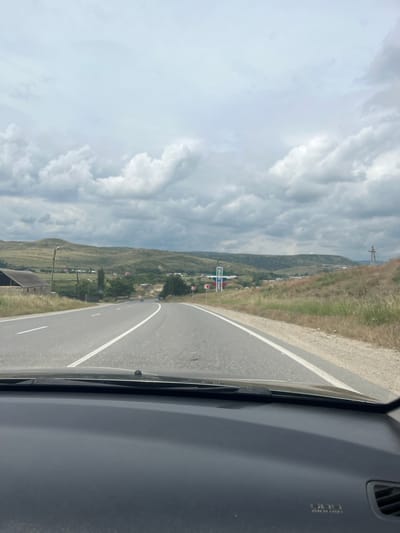 The image captures a view from inside a vehicle traveling on a paved road. The road is asphalt with distinct white lane markings. On the left side of the road, there is a building with a dark roof and stone walls, and a tall, thin structure resembling a lamppost or utility pole. The right side of the road features dry, yellowish grass and a gentle slope leading up to a grassy hillside dotted with bushes and small trees. In the distance, rolling green hills form the backdrop, with a smattering of houses and buildings visible in the valley. The sky is overcast with a layer of thick, gray clouds, suggesting a cloudy or potentially rainy day. There are signs visible on the side of the road, including what appears to be a directional sign with a red and white symbol and another sign with a blue and red design. A tall, cross-shaped structure, possibly a power line tower, stands on the right-hand hillside. The foreground shows the dark dashboard of the vehicle. The scene appears to be a rural or semi-rural area, likely in the vicinity of Karabudakhkent, Russia, given the context of the request.