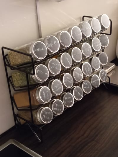 This image shows a black wire spice rack with four shelves filled with glass jars of spices. The jars have silver lids with round black labels written on in white chalk. The spice rack is positioned on a dark brown wooden countertop, likely in a kitchen. The background is a light-colored wall. The image is taken from a slightly elevated angle, looking down and across the spice rack. It is unclear if this image is located in Baden, Austria.