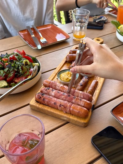 An outdoor dining scene at a restaurant in Byurakan, Armenia, features a wooden table laden with food and drinks. In the foreground, a partially filled glass with ice and a pink beverage sits to the left. To the right, a wooden board displays grilled sausages wrapped in bacon, a small bowl of mustard, and a small dish of what appears to be ketchup. A hand is using metal tongs to pick up one of the sausages. In the middle ground, a salad bowl filled with chopped tomatoes, cucumbers, peppers, and herbs is visible, along with a serving spoon. Another empty orange-colored plate with a fork and knife is to the left. In the background, two individuals are seated, one wearing a gray top and the other's arm with a ring is visible. A tall glass with a yellow beverage, keys, and a smartphone are also on the table. The overall setting suggests a meal being shared outdoors.