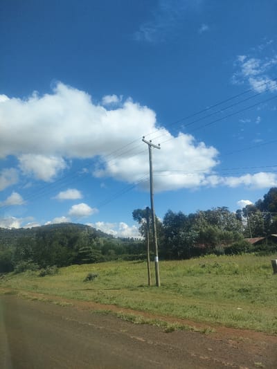 The image shows a rural landscape in Kibirichia ward, Kenya, under a partly cloudy sky. A utility pole with power lines stands in a grassy field. In the background are trees and a gently sloping hill. A road is visible in the foreground. A building or structure with a rusty roof is partly visible on the right.