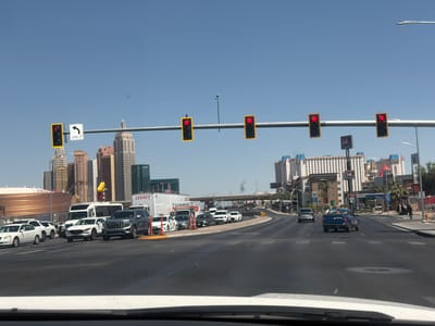 The image depicts a street scene in Paradise, Nevada, United States. The view is from inside a vehicle, looking out over the dashboard and windshield. The road stretches into the distance, with several cars driving on it. Overhead, a metal beam supports traffic lights, all displaying red signals. To the left, vehicles are stopped at the intersection. Buildings, including hotels and casinos, define the skyline, a mix of architectural styles visible against a clear, blue sky. A sign with a left turn arrow is visible to the side of the street at the edge of the image. The T-Mobile Arena is visible in the background.