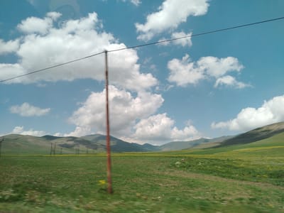 The image captures a wide, expansive view of a rural landscape under a bright, cloudy sky. In the foreground, a green, grassy field stretches out, with a few scattered yellow wildflowers visible. A thin, dark wire runs diagonally across the upper portion of the image, supported by a reddish-brown pole situated in the middle ground.
In the background, rolling green hills and mountains form a gentle backdrop. Several tall, lattice-like structures, likely power line towers, are sparsely dotted across the field and on the lower slopes of the hills, suggesting the presence of electrical infrastructure. The overall impression is one of open space and natural beauty, typical of a rural setting. The blurred motion in the foreground indicates the photo was likely taken from a moving vehicle.
