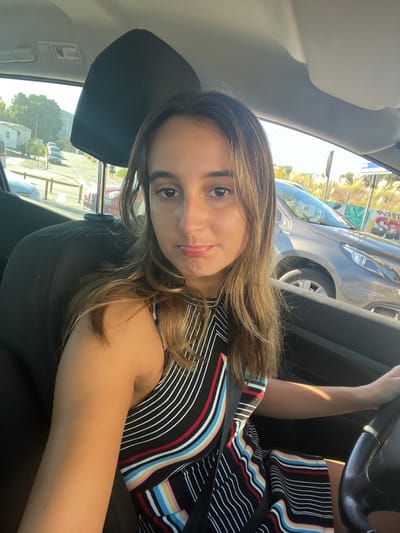 An adult female is seated in the driver's seat of a vehicle in Pedrouços, Portugal, capturing a self-portrait. She possesses light-toned skin, medium-length brown hair with lighter highlights, and is looking directly at the camera with slightly pouting lips. She is wearing a sleeveless black dress adorned with vertical and diagonal stripes in white, red, blue, yellow, and pink, secured by a dark seatbelt across her chest and lap. Her right hand is visible gripping the steering wheel.
The interior of the vehicle shows a dark headrest behind her and part of the dashboard. Outside the vehicle, on the left, a street ascends uphill. Several cars are visible, including a light-colored sedan and a red vehicle parked along the road, which is lined with white bollards. Further up the incline, trees and white modular structures are discernible. On the right side, a grey SUV or crossover is parked adjacent to the subject's car. Beyond this vehicle, a wall or structure featuring graffiti, including the letters "SC," is visible amidst sparse vegetation and under a bright, sunny sky.