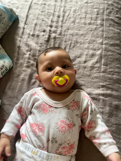 An infant is positioned centrally, lying supine on a textured, greyish-brown quilted surface, likely a blanket or bedspread, featuring a repeating pattern of small indentations. The infant has an olive to light brown complexion, dark hair, and a distinct dark mole on the upper left forehead. A yellow and pink pacifier is held in the infant's mouth. The infant's dark eyes are open and directed towards the camera. The infant is dressed in a white long-sleeved onesie adorned with a prominent pink rose floral pattern and white lower garment, possibly a diaper or soft pants. To the upper left of the frame, a segment of a blue and white patterned fabric object, possibly a pillow or folded garment with a floral motif, is visible. The scene is situated in Topoli, Bulgaria.