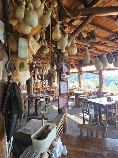 An indoor-outdoor dining establishment, located in the city of Tire, Turkey, is depicted under bright daylight. The scene is dominated by a rustic aesthetic, featuring a wooden beamed ceiling from which numerous pale, pear-shaped gourds, strings of dried red peppers, and bundles of dried herbs are suspended.
In the foreground, to the left, a dark vertical post supports a metal hook from which a string of dark beads and a dark fabric object hang. Several pieces of aged, crumpled paper are affixed to this post, along with a white electrical outlet. A dark wooden table occupies the lower right foreground, bearing a white salt shaker, a stand displaying a QR code, and clear drinking glasses. Adjacent to this table, a white plastic container filled with utensils rests on a beige fabric cover, next to a vintage-style green-bladed oscillating fan positioned on a wooden stand. The floor beneath the tables appears to be packed earth or gravel.
In the mid-ground, numerous light and dark stained wooden tables, mostly rectangular, are paired with matching wooden chairs. Along a central wooden post, three framed photographs are displayed vertically: a baby at the bottom, a young boy in the middle, and an adult's portrait at the top. A small, circular mirror is mounted between the top two frames.
In the background, the dining area opens to a panoramic view of dense green foliage and distant hills, illuminated by strong natural light entering from the right, which casts shadows across the floor. Several individuals are seated at tables in the background, appearing to be dining or conversing. The overall structure suggests an open-sided or semi-open air construction with a wooden roof and supporting pillars.