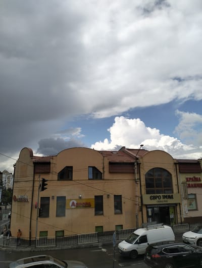 The image shows a street scene in Drohobych, Ukraine, with a prominent yellow building housing several businesses. The building features a curved facade, a tiled roof, and multiple windows of various shapes and sizes. Above the street level, there are signs for "МЕБЛІ ДИВОНОРА" (furniture store), "АВРОРА МУЛЬТИМАРКЕТ" (supermarket), and "ЄВРО ТРЕНД СЕКОНД ХЕНД" (second-hand clothing store). A traffic light is visible on a pole in front of the building.
The sky is partly cloudy, with large, dramatic grey storm clouds dominating the upper left portion of the frame and bright white cumulus clouds against a blue sky in the upper right.
Several people are visible on the sidewalk in front of the building, walking and standing. There are also cars on the street, including a white van with a roof rack, a black SUV, and a white sedan. The street appears wet, suggesting recent rain. The overall impression is of a moderately busy urban environment under a dynamic sky.